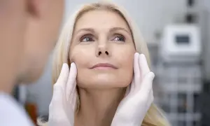 Manos enguantadas evaluando el rostro de una mujer rubia, preparándose para un tratamiento de fotorejuvenecimiento facial.