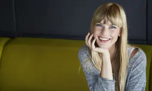 Sonrisa radiante de una joven rubia con flequillo, apoyando su barbilla en la mano. ¡Se ve genial al probar flequillo!