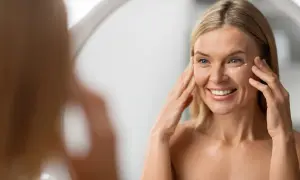Mujer sonríe frente al espejo, aplicándose crema en el contorno de ojos para atenuar las líneas de expresión.