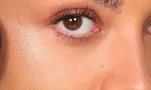Primer rostro con brocha negra aplicando producto no comedogénico. Piel radiante y ojos marrones con sombras rosadas.