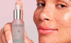 Una mano sostiene el Agua de Belleza Caudalie, mientras una mujer con pecas sonríe. Opiniones positivas sobre este elixir para una piel radiante.