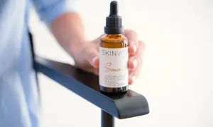 Mano sostiene un frasco de vidrio ámbar con gotero, etiquetado como "SKINVITY care Serum". Este booster es un sérum facial.