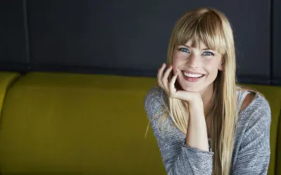 Sonrisa radiante de una joven rubia con flequillo, apoyando su barbilla en la mano. ¡Se ve genial al probar flequillo!