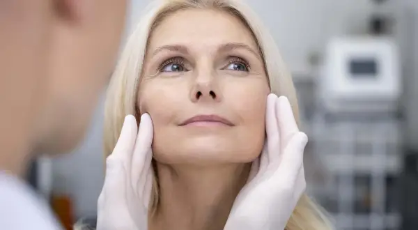 Manos enguantadas evaluando el rostro de una mujer rubia, preparándose para un tratamiento de fotorejuvenecimiento facial.