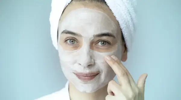 Mujer con toalla en la cabeza y mascarilla blanca en el rostro, aplicándose un peeling facial.