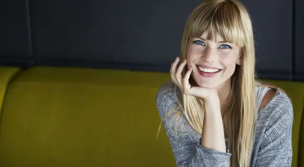 Sonrisa radiante de una joven rubia con flequillo, apoyando su barbilla en la mano. ¡Se ve genial al probar flequillo!