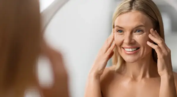 Mujer sonríe frente al espejo, aplicándose crema en el contorno de ojos para atenuar las líneas de expresión.
