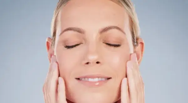 Mujer con ojos cerrados y una sonrisa suave, acariciando su rostro para realzar su óvalo facial.
