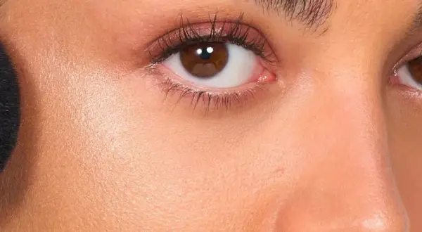 Primer rostro con brocha negra aplicando producto no comedogénico. Piel radiante y ojos marrones con sombras rosadas.