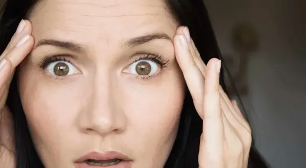 Mujer con ojos muy abiertos y arrugas en la frente, tocándose las sienes con las manos.