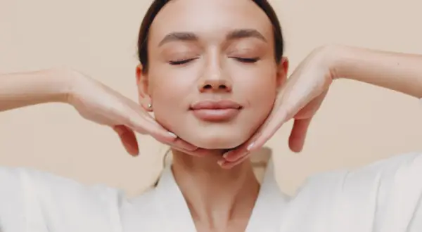 Mujer con ojos cerrados, disfrutando de un masaje facial. Sus manos sostienen suavemente su rostro, practicando yoga facial para una piel radiante.