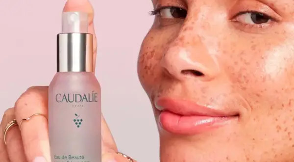 Una mano sostiene el Agua de Belleza Caudalie, mientras una mujer con pecas sonríe. Opiniones positivas sobre este elixir para una piel radiante.