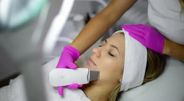 Una esteticista realiza un peeling ultrasonico a una mujer. El dispositivo emite vibraciones para limpiar y rejuvenecer la piel.