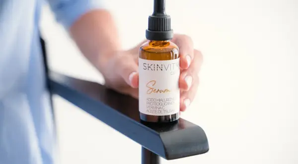 Mano sostiene un frasco de vidrio ámbar con gotero, etiquetado como "SKINVITY care Serum". Este booster es un sérum facial.