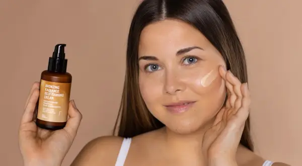 Mujer aplica bronceador freshly en su mejilla. Sostiene un frasco de crema autobronceadora.