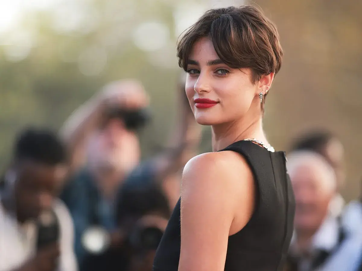 Mujer con un moderno corte bixie, labios rojos y mirada intensa, rodeada de fotógrafos.