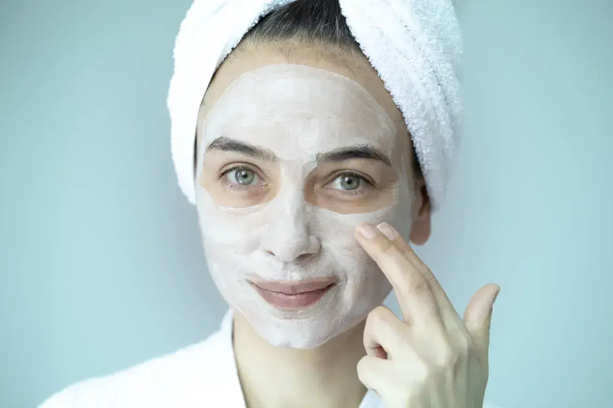 Mujer con toalla en la cabeza y mascarilla blanca en el rostro, aplicándose un peeling facial.