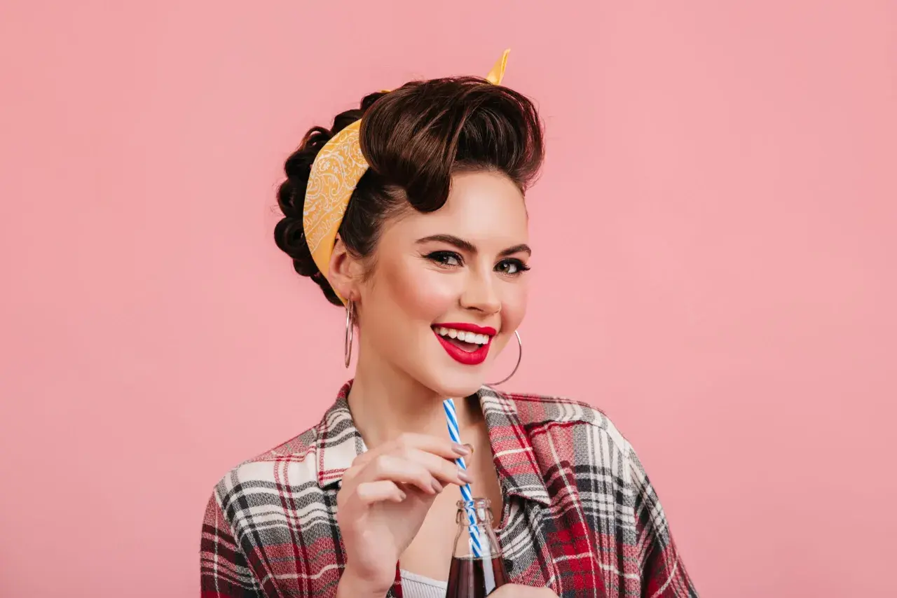 Mujer sonriente con peinados vintage y pañuelo amarillo, bebiendo de una botella.
