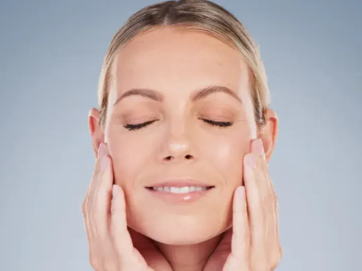 Mujer con ojos cerrados y una sonrisa suave, acariciando su rostro para realzar su óvalo facial.