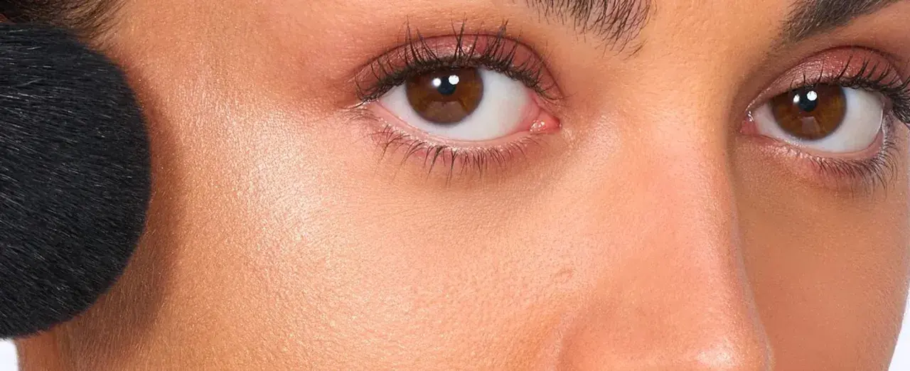 Primer rostro con brocha negra aplicando producto no comedogénico. Piel radiante y ojos marrones con sombras rosadas.