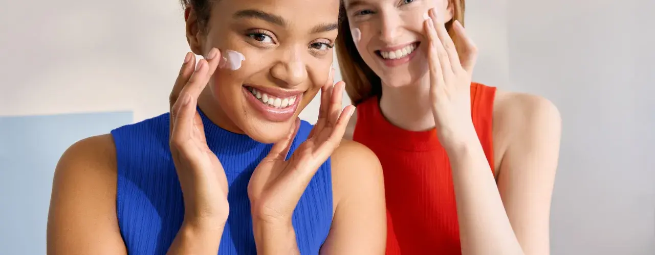 Dos mujeres sonríen mientras se aplican crema en el rostro. Ambas tienen piel normal y lucen radiantes.