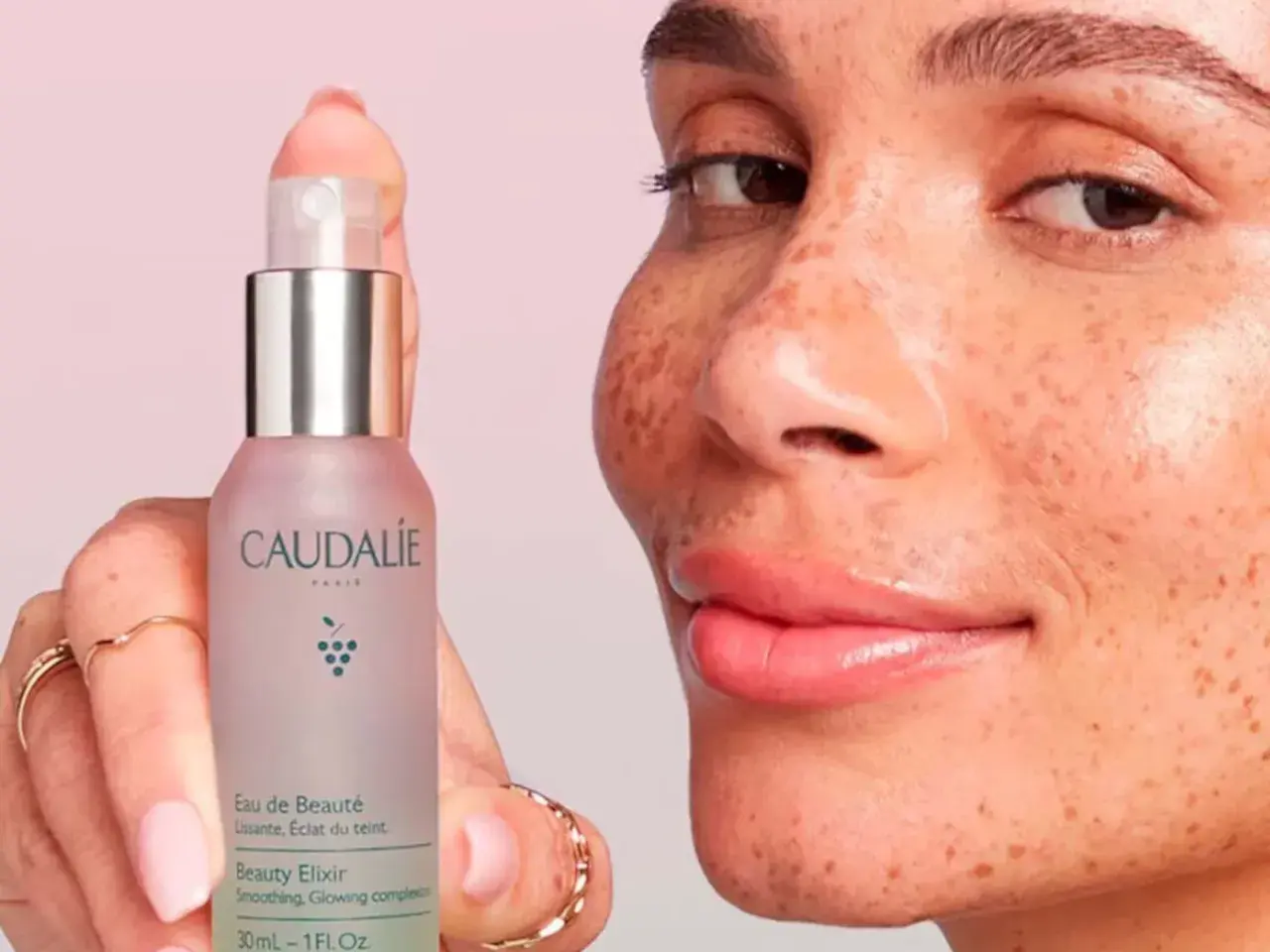 Una mano sostiene el Agua de Belleza Caudalie, mientras una mujer con pecas sonríe. Opiniones positivas sobre este elixir para una piel radiante.