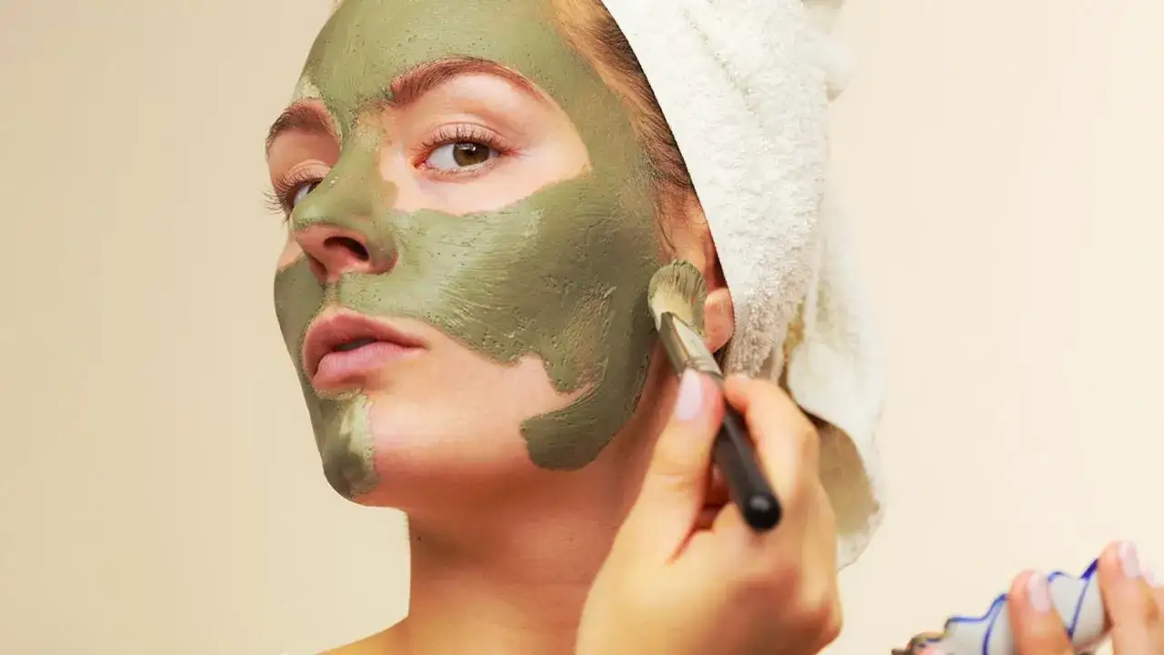 Mujer aplic&aacute;ndose mascarilla de arcilla verde en el rostro con brocha. La arcilla verde sirve para limpiar y purificar la piel.