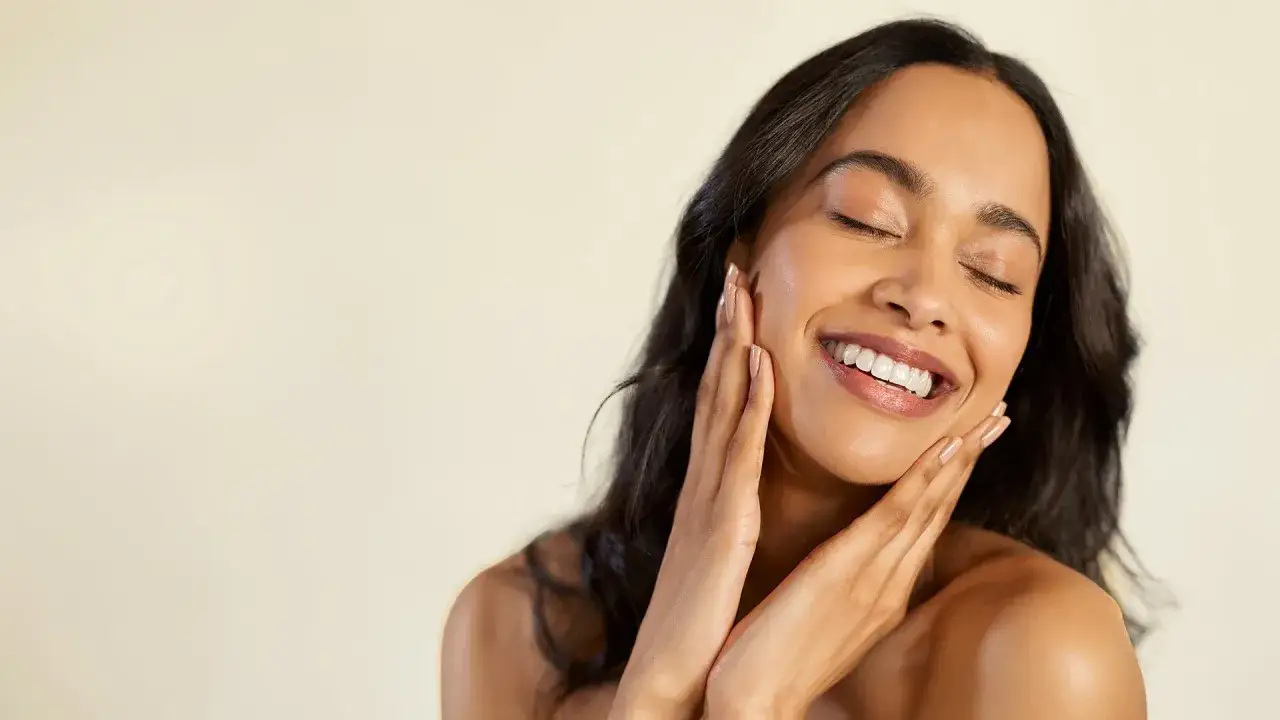 Mujer sonríe con los ojos cerrados, acariciando su rostro. Su piel normal luce radiante y saludable.