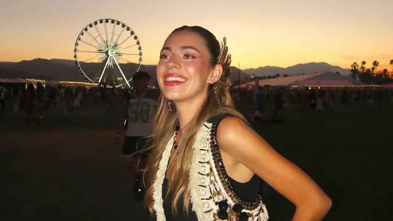 Joven sonriente con maquillaje festival, adornos en el cabello y chaleco bordado, disfrutando del atardecer en un evento con noria y multitudes.