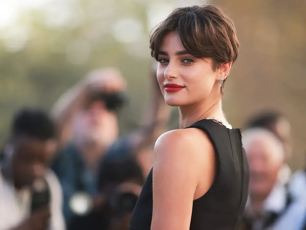 Mujer con un moderno corte bixie, labios rojos y vestido negro, rodeada de fot&oacute;grafos.