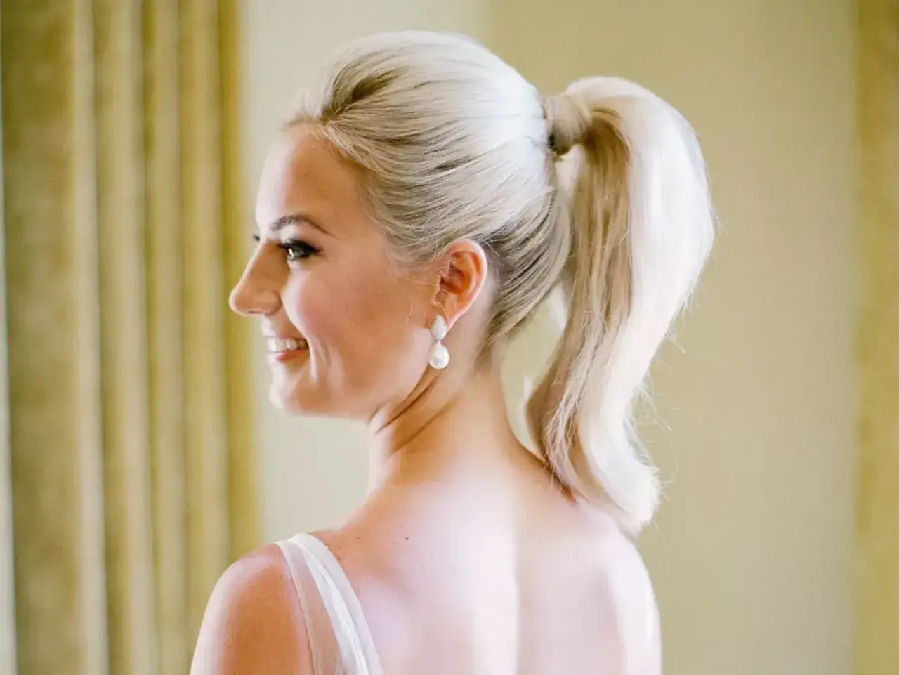 Novia sonriente con un elegante peinado coleta alta boda, luciendo pendientes de perla.