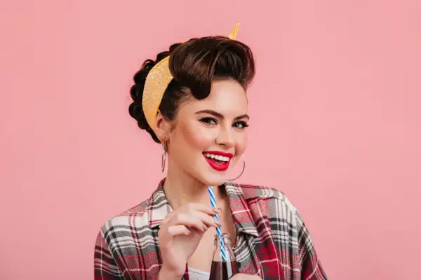 Mujer sonriente con peinados vintage y pañuelo amarillo, bebiendo de una botella.