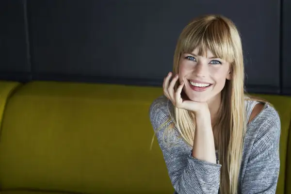 Sonrisa radiante de una joven rubia con flequillo, apoyando su barbilla en la mano. ¡Se ve genial al probar flequillo!