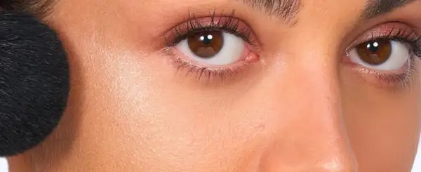 Primer rostro con brocha negra aplicando producto no comedogénico. Piel radiante y ojos marrones con sombras rosadas.