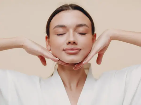 Mujer con ojos cerrados, disfrutando de un masaje facial. Sus manos sostienen suavemente su rostro, practicando yoga facial para una piel radiante.