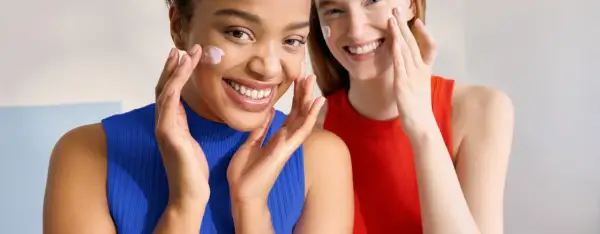 Dos mujeres sonríen mientras se aplican crema en el rostro. Ambas tienen piel normal y lucen radiantes.