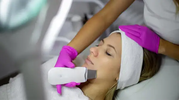 Una esteticista realiza un peeling ultrasonico a una mujer. El dispositivo emite vibraciones para limpiar y rejuvenecer la piel.